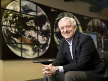 David Titley, Penn State alum, retired U.S. Navy rear admiral sitting on a chair  with hands clasped with electronic wall depicting full earth globes behind him.