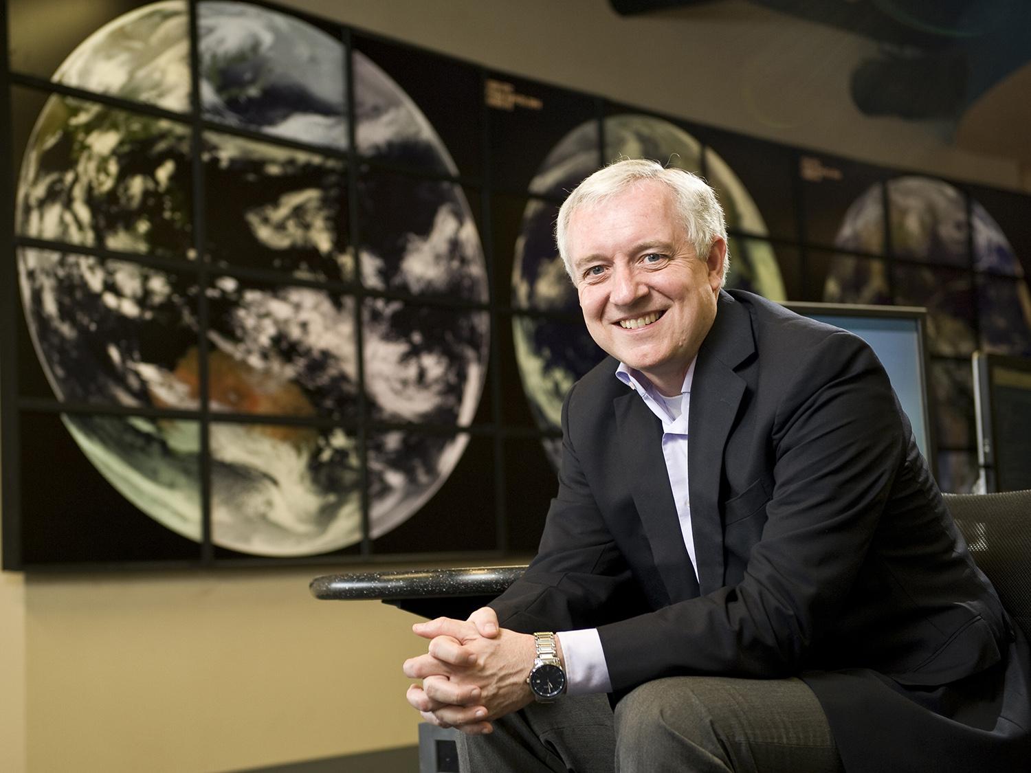 David Titley, Penn State alum, retired U.S. Navy rear admiral sitting on a chair  with hands clasped with electronic wall depicting full earth globes behind him.